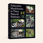 A hardcover book titled 'Psilocybin Mushrooms in Their Natural Habitats' by Paul Stamets, featuring a black cover with multiple color photographs of mushrooms and white text, shown on a light beige background.