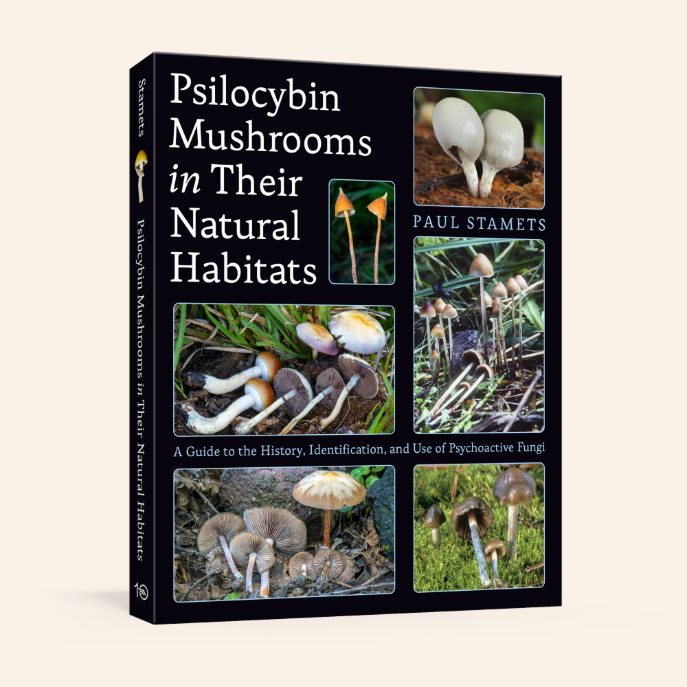A hardcover book titled 'Psilocybin Mushrooms in Their Natural Habitats' by Paul Stamets, featuring a black cover with multiple color photographs of mushrooms and white text, shown on a light beige background.