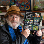 Paul Stamets holding his book titled 'Psilocybin Mushrooms in Their Natural Habitats' in a colorful indoor setting.