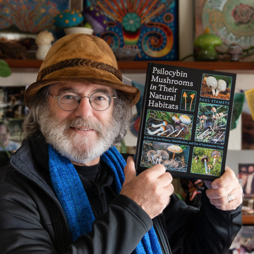 Paul Stamets holding his book titled 'Psilocybin Mushrooms in Their Natural Habitats' in a colorful indoor setting.